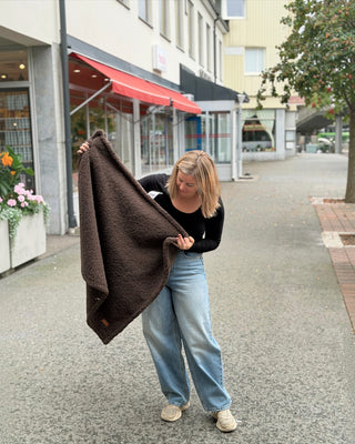 Kvinna från Hundinspiration håller upp en mjuk och fluffig brun CAZO Mirson hundfäll utomhus på en stadsgata.