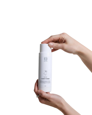 Hundinspiration Pennys Signature Conditioner i en vit flaska som hålls av två händer mot en vit bakgrund.